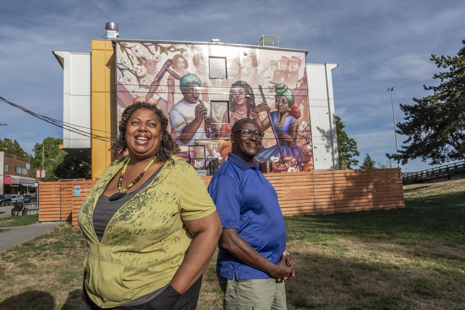 A smiling Black couple standing in a grassy area with a mural of black artists and musicians on a building behind them